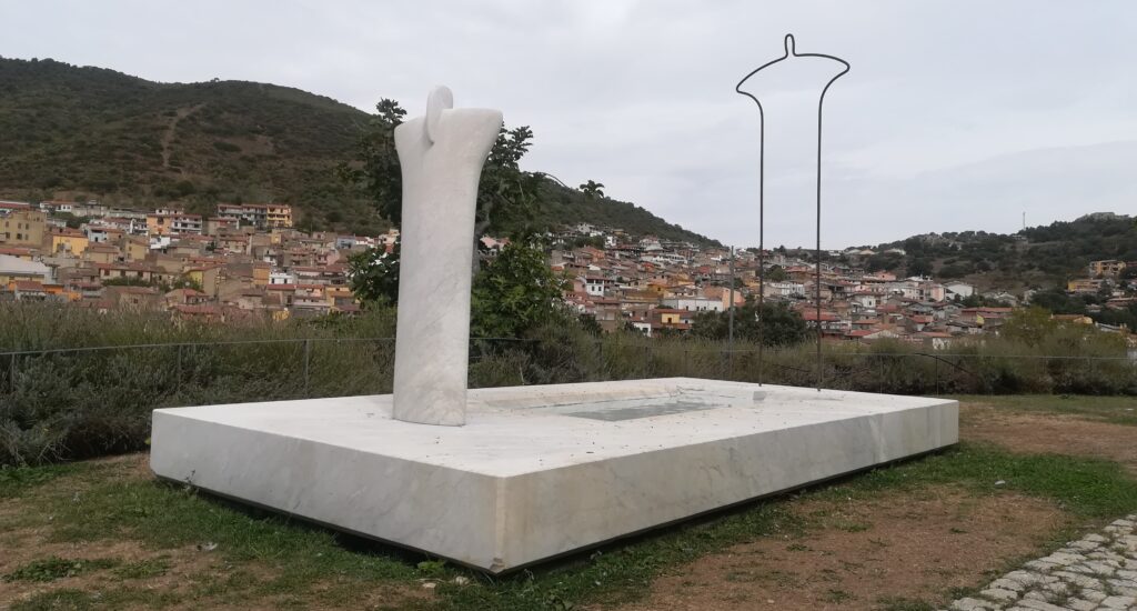 La Madre
Scultura monumentale in marmo di Carrara. Sotto un parallelepipedo rettangolare sopra la statua che rappresenta con le sue forme semplici, lineari la Dea Madre. La figura si allarga verso l'alto. Dietro, sullo sfondo, si arrampica il paese di Orani