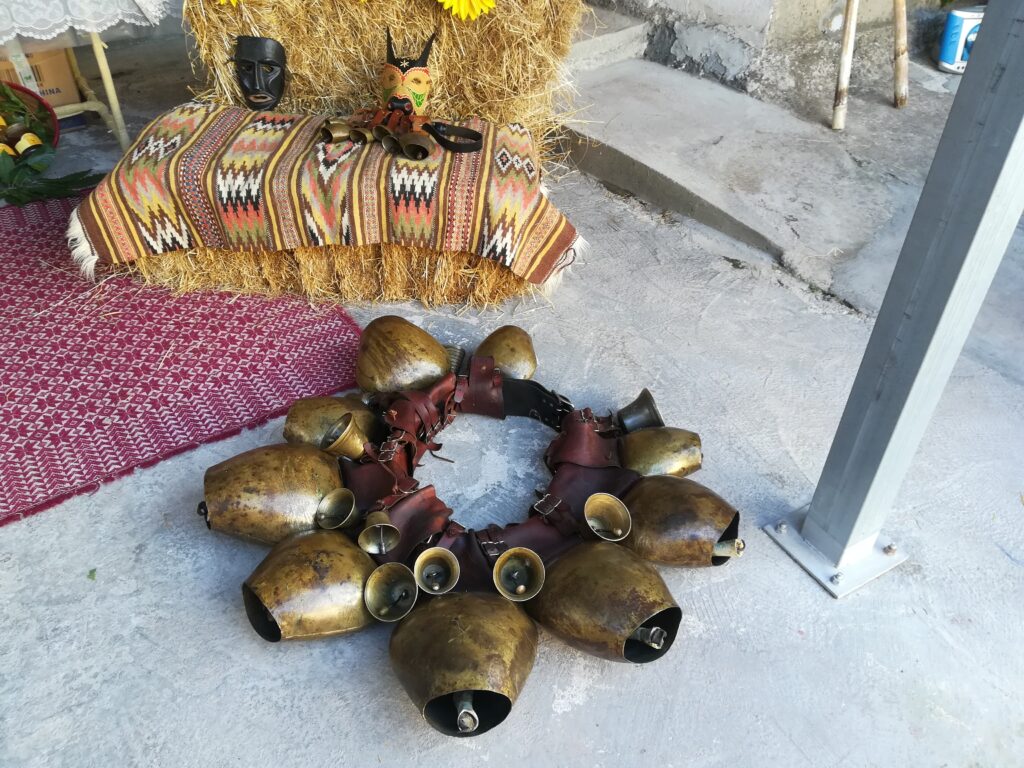 Per terra circolo di campanacci, dietro, sopra una balla di fieno coperta da un tappeto a fiamme di Nule, machere del Carnevale.