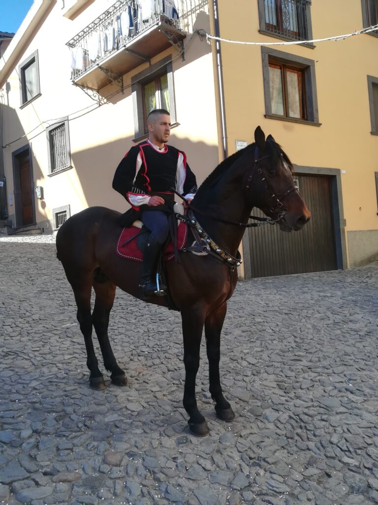Sopra un cavallo addobato solo con collare colorato, il cavaliere ancora senza la maschera, con abito nero in velluto e strisce rosse.