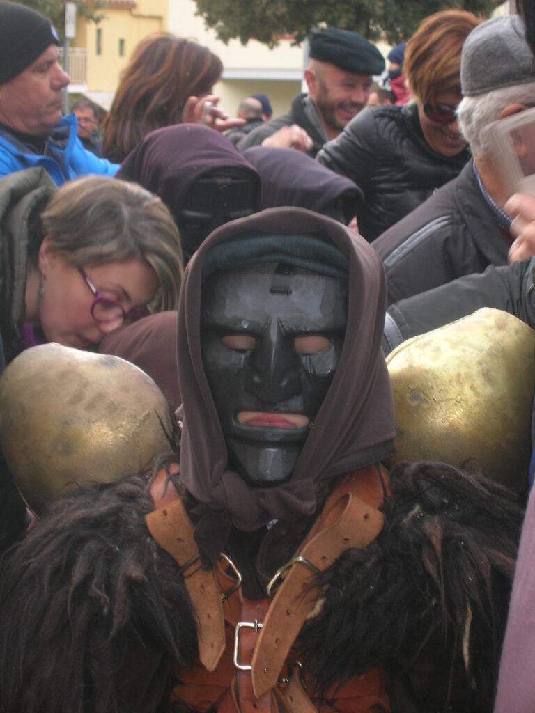 In primo piano il Mamuthone, la sua maschera di legno nera con fazzoletto marrone sopra a incorniciare il viso, la pelle come giacca nera e le fibbie di cuoio che reggono i campanacci dietro la schiena.