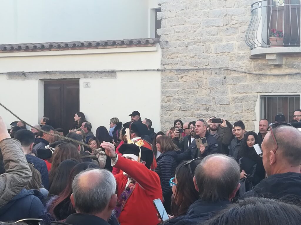 Mamoiada, nelle vie tante persone con al centro un uomo con vestito rosso che lancia una fune.