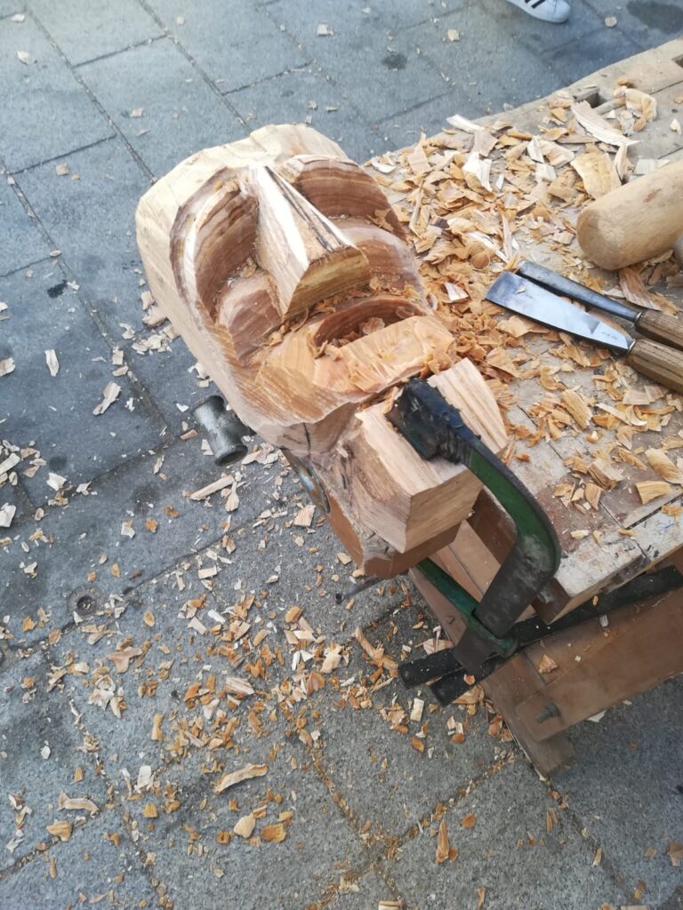 Sopra un banchetto di legno, un pezzo di legno viene intagliato abib
lmente per creare una maschera.