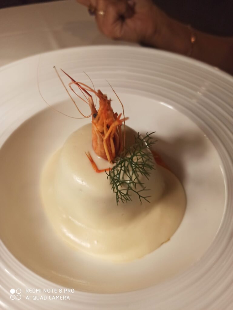 Al centro di un grande piatto di ceramica lucida bianco con tesa molto larga, troviamo adagiato un piccolo zucchettone di melanzane ricoperte dalla salsa al pecorino bianca, con sopra la testa del gambero e una foglia di aneto