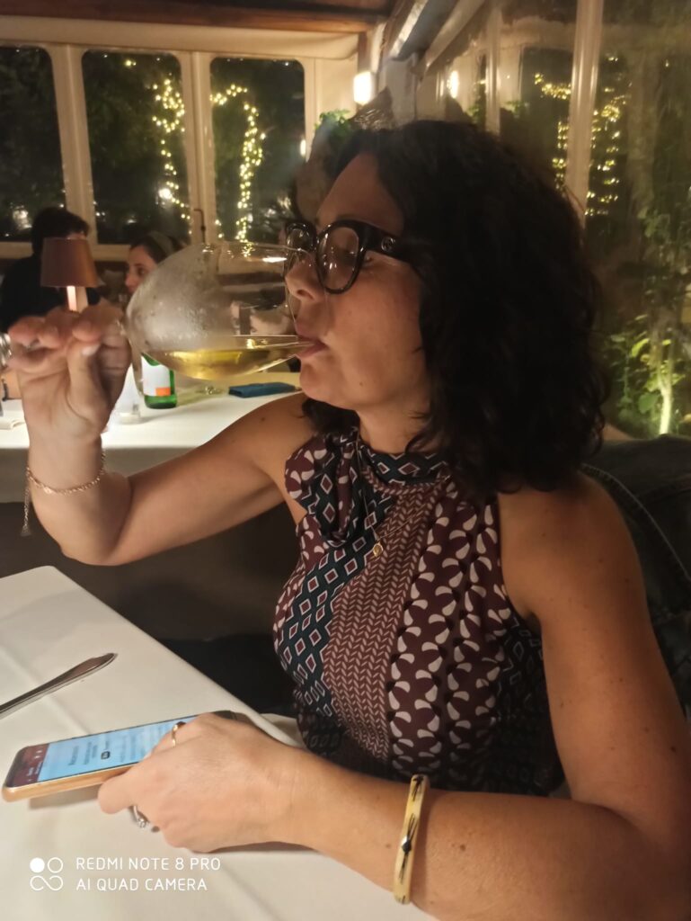 Seduta al tavolo del ristorante mentre degusto un calice di vino bianco. Indosso una camicia di colori blu e bordeaux all'americana, con fiocco sul collo.